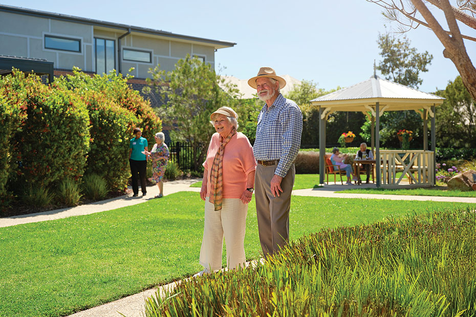 Residents walking and relaxing in the lush gardens of Benetas Corowa Court, with a gazebo and well-maintained greenery in the background.