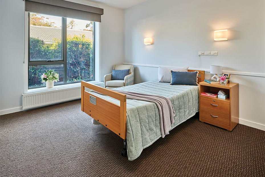 A bright and comfortable resident room at Benetas Corowa Court, featuring a large window, a single bed, a cosy chair, and a wooden bedside table with soft lighting.