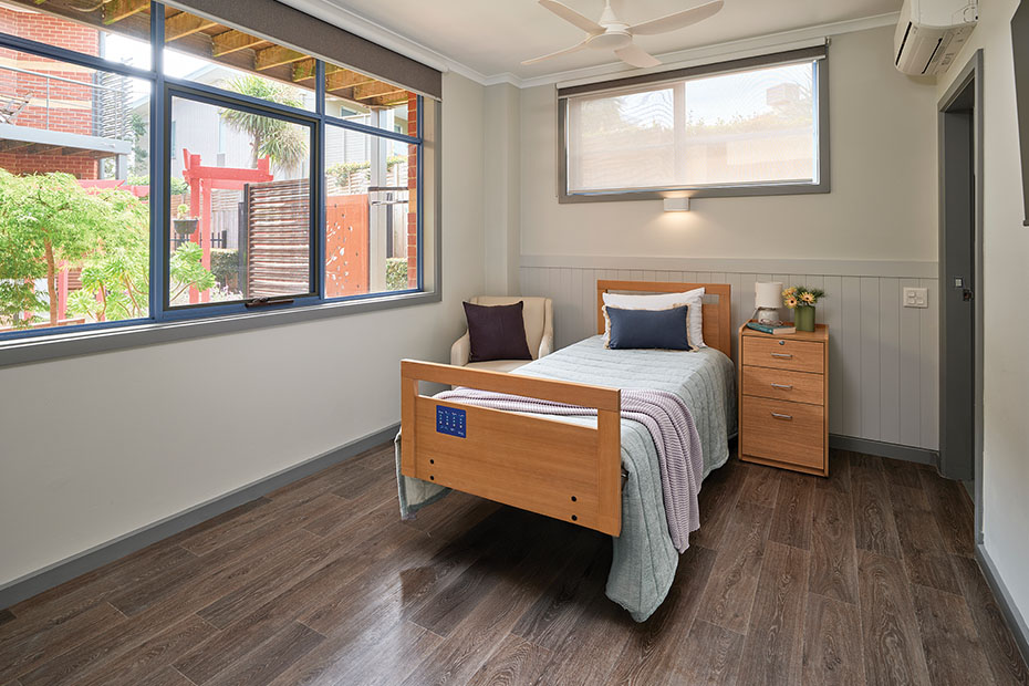 A modern and spacious resident room at Benetas Corowa Court, with a large window providing a view of the garden, a single bed, a ceiling fan, and sleek wooden flooring.