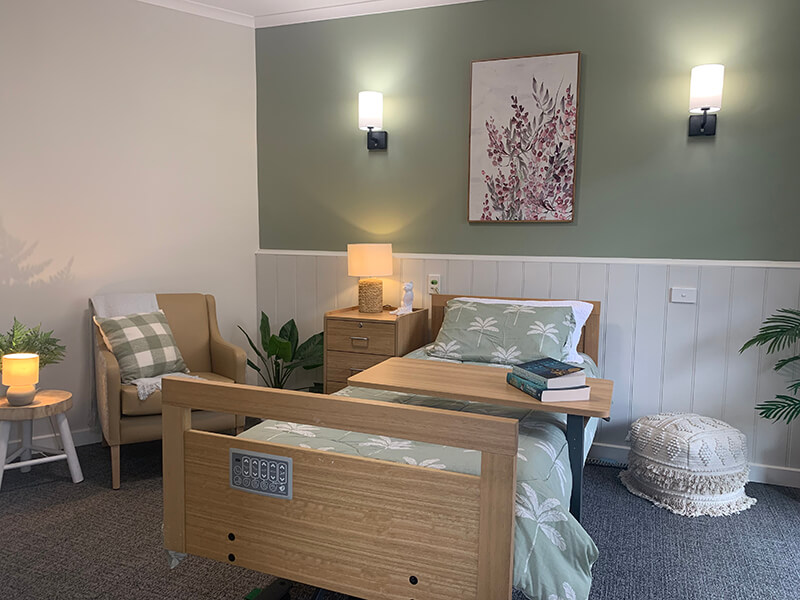 Modern aged care bedroom with a single bed, armchair, bedside table, soft lighting, and calming green decor at Benetas.