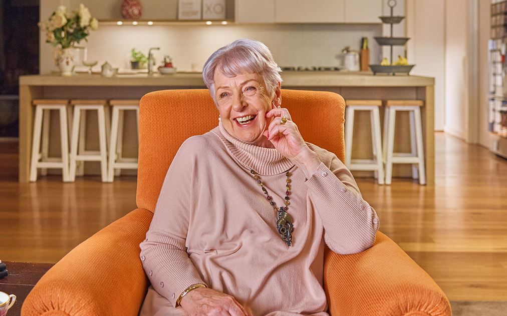 An older woman smiles while seated in a vibrant orange chair, exuding warmth and joy in her expression.