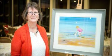 A woman in an orange sweater stands beside a painting, adding a vibrant touch to the artwork.