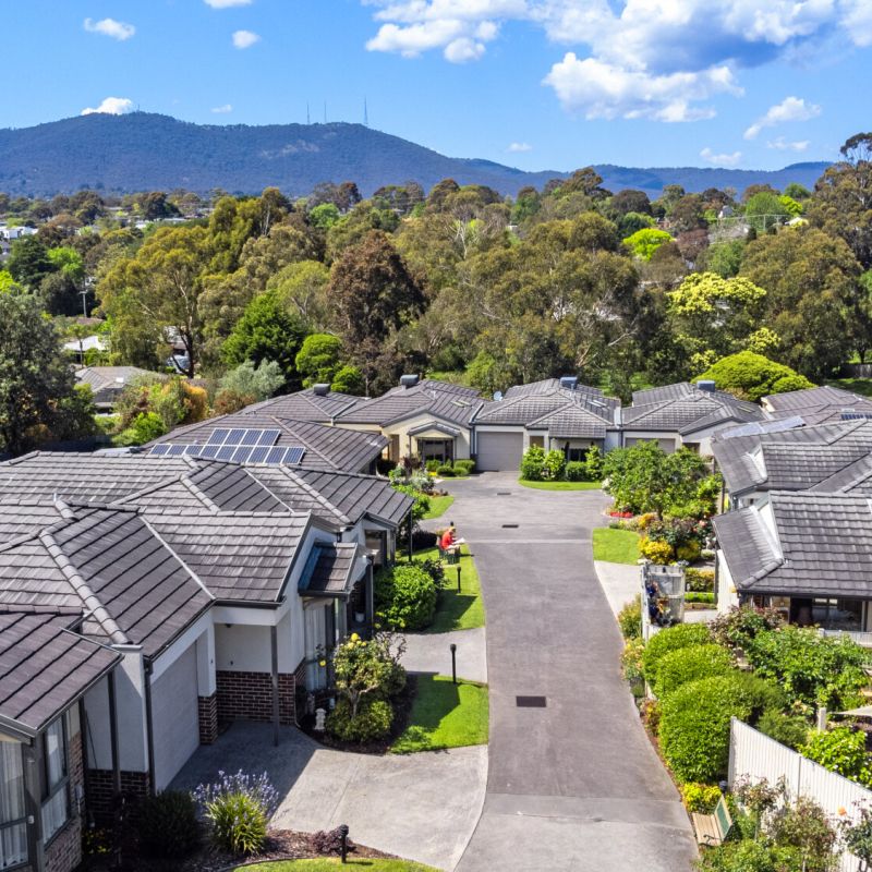 Aerial view of a modern retirement community with landscaped gardens, tiled villa rooftops and surrounding leafy suburban streets set against a mountain backdrop